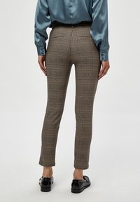 Personne portant un pantalon à carreaux ajusté, un chemisier en satin teal et des mocassins noirs, se tenant contre un fond clair uni, de dos.