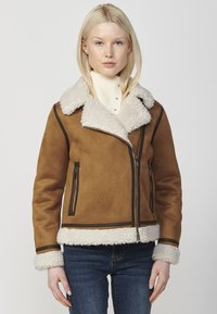 Giubbotto in shearling marrone con colletto in morbido pile, chiusura con zip e tasche, rifiniture scure a contrasto e finitura in tessuto testurizzato.