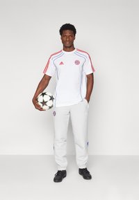 Biała koszulka Adidas z czerwonymi i niebieskimi akcentami, szare spodnie, czarne buty oraz czarno-biała piłka nożna. Na koszulce znajduje się logo klubu.