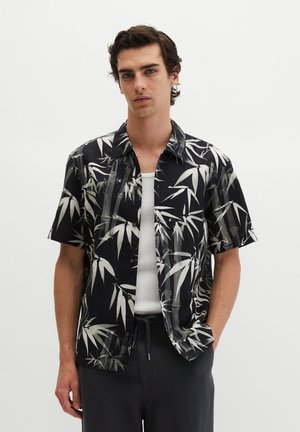 Młody mężczyzna w czarnej krótkiej koszuli zapinanej na guziki z białym nadrukiem bambusa na białym tank topie, ręce w ciemnych kieszeniach spodni.