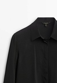 Chemise noire à manches longues avec col pointu et patte de boutonnage dissimulée, suspendue sur un fond blanc.