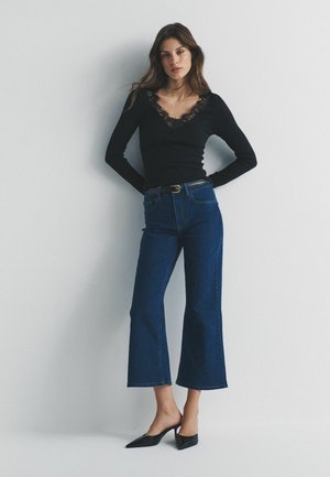 Sieviete stāv pie vienkāršas sienas, valkājot melnu blūzi ar mežģīņu apmali, tumši zilas plaša stila džinsa bikses, melnu jostu un melnus kurpes ar smailiem purngaliem.