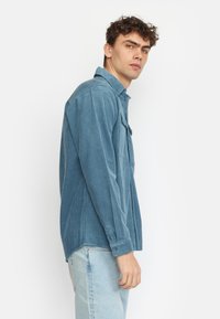 Camicia in velluto corduroy blu con maniche lunghe, polsini con bottoni e colletto. Presenta coste verticali e una tasca sul petto. Indossata con jeans chiari.