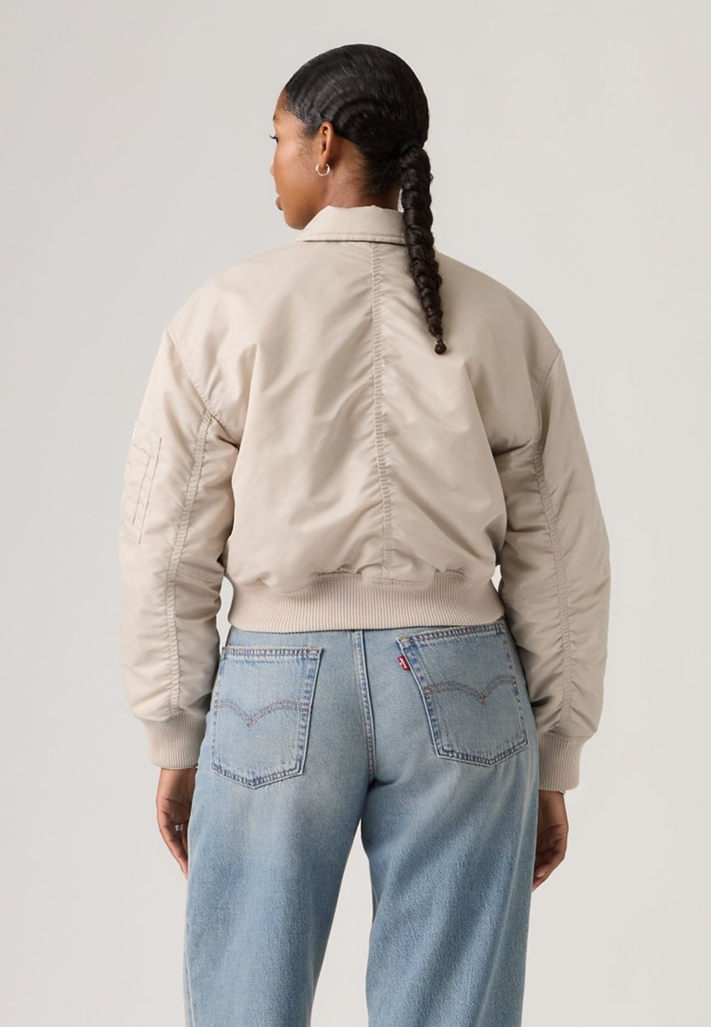 Femme aux cheveux tressés portant une veste aviateur beige et un jean taille haute bleu clair, de dos, sur un fond uni.