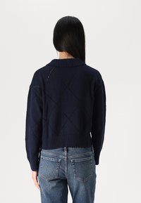 Mornarskoplavseni pleteni džemper s dijamantnim uzorkom, rebrastim rubom i ovratnikom, skraćene dužine, uparen s svijetloplavim trapericama.