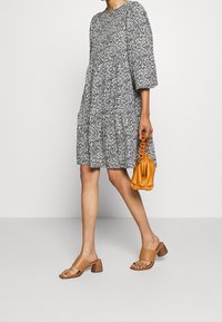 Robe à imprimé floral bleu marine et blanc, coupe ample, manches ballons, design à étages. Assortie avec des sandales à glisser beiges et un sac à main orange.