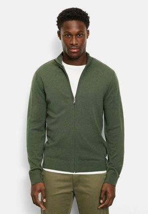 Maglione verde con zip, in tessuto a coste e polsini. Indossato sopra una maglietta bianca, presenta un colletto alto e una cerniera liscia.