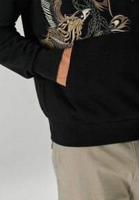 Hanorac negru cu broderie complexă multicoloră, având un design cu fenix. Include un buzunar frontal și manșete și tiv cu dungă.
