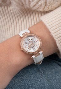 Käekell valge nahkraadi, roosakuldse korpuse ja kristallidega, ning valgel kellal kristallidest lillekujundiga, kantud randmel.