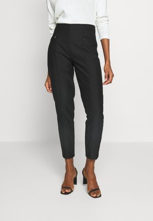 Femme portant un pantalon noir taille haute fuselé avec poches avant zippées et des sandales à talons ouvertes noires, debout devant un fond uni.