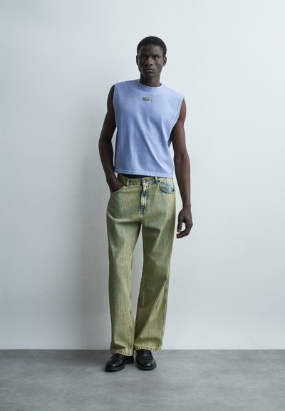 Άνδρας που φορά αμάνικο ανοιχτό μπλε τοπ και φαρδιά ξεθωριασμένα πράσινα τζιν, στέκεται μπροστά σε έναν ανοιχτό τοίχο με τα χέρια στις τσέπες.