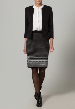 Frau trägt weiße Bluse, schwarzes kurzes Blazer, gemusterten schwarz-weißen Bleistiftrock, schwarze Strumpfhosen und schwarze Stiefeletten, steht.