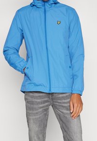 Light blue zip-up jacket with a hood, elastic cuffs, and two side pockets. Features a small yellow logo on the left chest area.