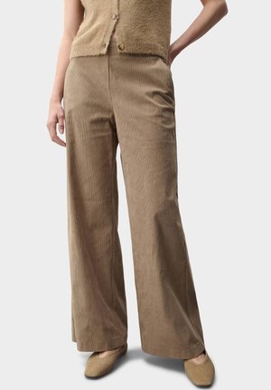 Vrouw draagt hoge taille, wijde pijpen, bruine corduroy broek, beige zachte geknoopte vest en beige platte schoenen tegen een effen achtergrond.