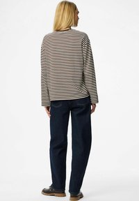 Žena s dugom plavom kosom stoji, obučena u smešnu smešnu majicu dugih rukava smeđe i bijele pruge, tamnoplave traperice i crne cipele s smešnim smešnim smešnim smešnim smešnim smešnim smešnim smešnim smešnim smešnim smešnim smešnim smešnim smešnim smešnim smešnim smešnim izlaskom.