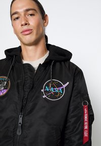 Zwarte bomberjack met capuchon, voorzien van geborduurde NASA- en Apollo-patches, trekkoorden en een rode "Remove Before Flight"-label.
