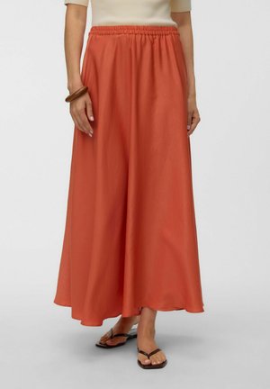 Person iført en lang orange nederdel, beige kortærmet top, brun armbånd og mørke klipklapper, stående mod en ensfarvet baggrund.
