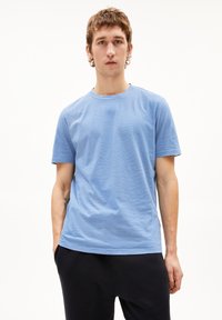 Junger Mann mit kurzen braunen Haaren, der ein hellblau gestreiftes T-Shirt und schwarze Hosen trägt, steht vor einem weißen Hintergrund.