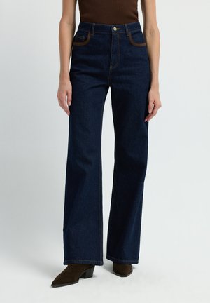 Brede pijpen van donker denim met contrasterende bruine stiksels, voorzakken en een hoge taille; gecombineerd met bruine hakken.