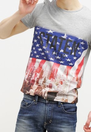 Person iført en grå T-shirt med et amerikansk flag, bybillede og ordet "STATEN" trykt på, kombineret med blå jeans.