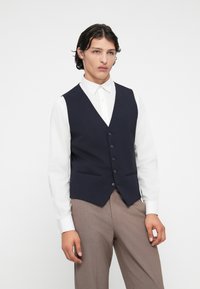 Colete azul marinho com decote em V, fechamento de cinco botões e dois bolsos, combinado com uma camisa branca e calças castanhas claras. Textura de tecido suave.