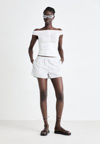 Weißes schulterfreies Oberteil mit Raffung, gestreifte hellgraue Shorts und schwarze flache Sandalen mit Knöchelriemen. Minimalistisches Design.