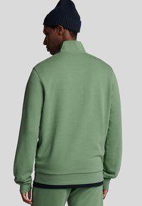 Sudadera de forro polar verde con cuello alto, mangas largas y puños acanalados. Combinada con una gorra de lana azul marino, que presenta una textura suave y un diseño minimalista.