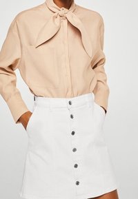 Blouse beige avec un col noué et une poche, associée à une jupe en denim blanche dotée d'une fermeture à boutons et d'un ourlet droit.
