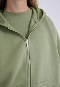 Felpa con zip verde chiaro dal tocco morbido, con tasca frontale e cappuccio. Indossata sopra una maglietta a maniche corte coordinata.