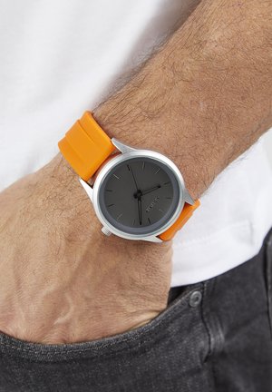 Silver wristwatch with a round, black face, orange silicone strap, and minimalistic index markers. The band features a smooth texture and modern design.