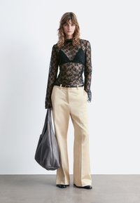 Schwarzes Oberteil mit langen Spitzenärmeln, das über einem schwarzen BH getragen wird, kombiniert mit beigen, hochgeschnittenen, weit geschnittenen Hosen. Graue Ledertasche. Schwarze Schuhe.