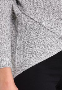 Gros plan d'une personne portant un pull en maille gris clair avec un ourlet diagonal et un pantalon noir, montrant une partie du bras et du torse.