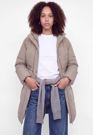 Une femme se tient devant un fond blanc. Elle porte un manteau matelassé couleur taupe, une chemise blanche et un jean bleu, avec un pull gris noué autour de la taille.