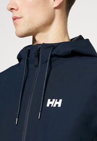 Chaqueta con capucha navy hecha de una tela suave; cuenta con un cierre frontal, cordones ajustables y un logotipo blanco en el pecho.