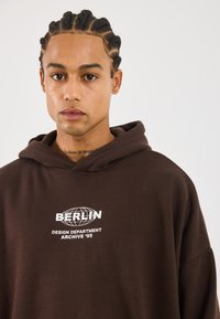 Bruine hoodie met witte bedrukte tekst "BERLIJN" en "DESIGN DEPARTMENT ARCHIVE '95." Heeft een relaxed fit en een zachte structuur.