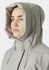 Chaqueta impermeable de color gris claro con una gran capucha, cierre de cremallera y cuerdas ajustables. Presenta una textura suave y un diseño minimalista.