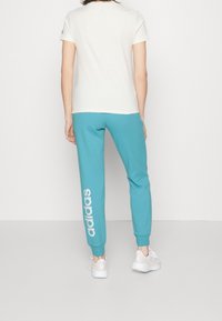 Person wearing turquoise Adidas sweatpants with white logo on left leg, white short-sleeve shirt, and white athletic shoes standing facing away.