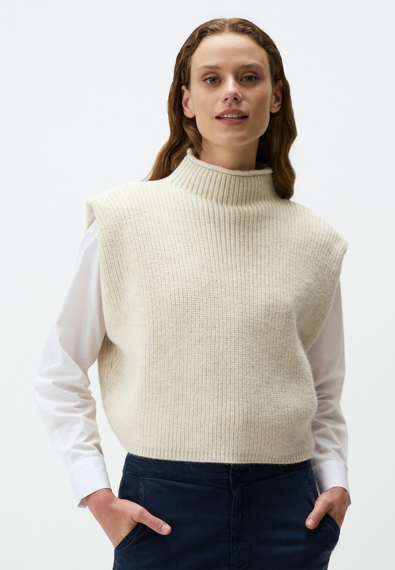 Pull en maille sans manches couleur crème avec un col haut côtelé et un tissu texturé, superposé à une chemise blanche à col.