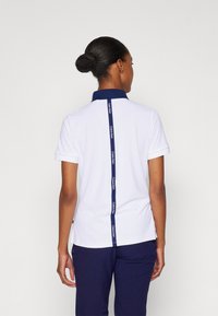 Bílá polo košile s námořnickým límečkem, krátkými rukávy a vertikálním námořnickým logem podél zad. Střih je přiléhavý.