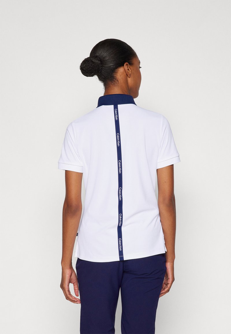 Bílá polo košile s námořnickým límečkem, krátkými rukávy a vertikálním námořnickým logem podél zad. Střih je přiléhavý.