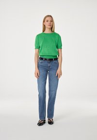 Vrouw staat in een groene trui met korte mouwen, blauwe jeans met een zwarte riem en zwarte studsloffen op een witte achtergrond.