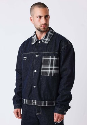 Homme aux cheveux courts portant une veste en jean foncée avec un col, une poche et un ourlet à carreaux, debout devant un fond clair uni.