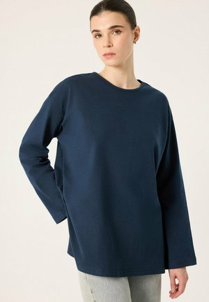 Femme portant une chemise ample bleu foncé à manches longues et un pantalon gris clair, debout avec une main sur la hanche devant un fond uni.