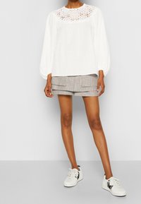 Hvid langærmet bluse med blonde detaljer ved halsudskæringen, parret med ternede shorts og hvide sneakers med sorte accenter.