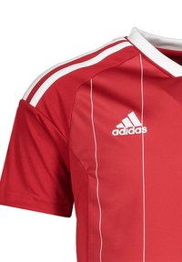 Maillot de sport rouge en tissu léger, avec des rayures blanches sur les épaules et des accents verticaux, comportant un logo brodé.