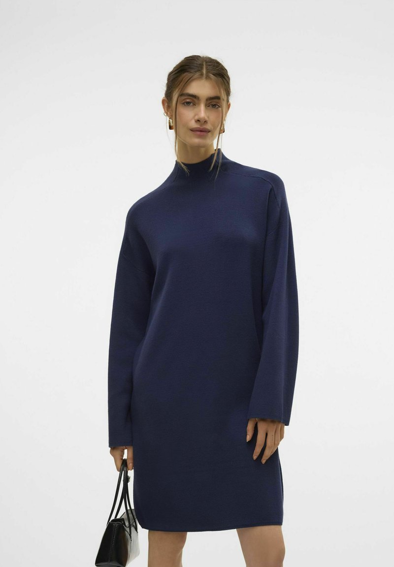 Abito blu navy con maniche lunghe e collo alto, realizzato in un tessuto strutturato. Abbinato a una piccola borsa nera. Silhouette semplice e dritta.