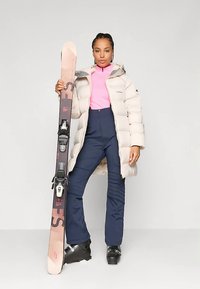 Γυναίκα σε μπεζ μπουφάν, ροζ μπλούζα, ναυτικές σκιέρ παντελόνι και μαύρες σκιερικές μπότες κρατά ένα ζευγάρι ροζ και μαύρα σκι όρθια.