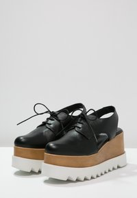 Chaussures à plateforme en cuir noir avec un design découpé, semelle intermédiaire en bois brun et semelle extérieure en caoutchouc blanc à bords dentelés. Présente un système de laçage.