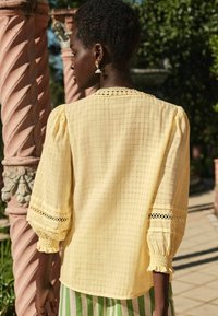 Camicia gialla a scacchi con maniche a palloncino, dettagli in pizzo sui polsini e scollatura arricciata. Tessuto leggero e texturizzato.