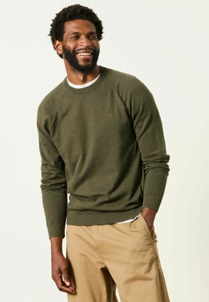 Mand smiler iført en olivengrøn sweater og beige bukser med den ene hånd i lommen mod en ensfarvet lys baggrund.